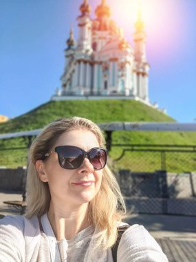 A young woman with glasses takes a walk in the historical place of Kyiv - Andreevsky Descent. Church in the background on a sunny day. Ukraine, Europe clipart