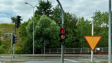 Geniş otoyol, bulutlu bir gökyüzünün altında giden arabalarla yeşil bir manzara boyunca uzanır. Sahne yemyeşil ağaçlar ve mavi korkuluklar tarafından çerçevelenmiş, huzurlu bir yol yolculuğunu yakalıyor..