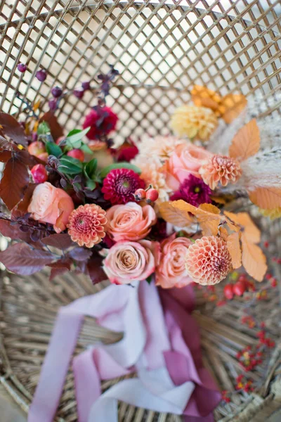 stock image Beautiful autumn wedding bouquet with orange and red flowers and berries. Autumn bouquet with ribbons on a rattan peacock chair