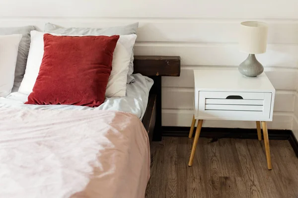 stock image Bedroom interior with bed and table. Bed with white and red pillows, pink blanket and small bed table with lamp near wooden white wall