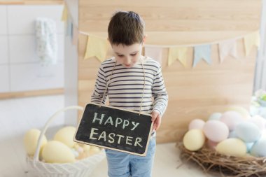 Paskalya süsleriyle donatılmış hafif bir mutfağın içinde paskalya tabelası olan beyaz bir çocuk elinde karatahta tutuyor.