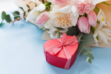 Beautiful bouquet with roses, carnations, iris and eucalyptus and red heart gift box on a blue background, copy space. Mothers day, 8 march, holiday gift concept