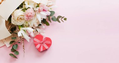 Beautiful bouquet with roses, carnations, iris and eucalyptus and red heart gift box on a pink background, copy space. Mothers day, 8 march, holiday gift concept