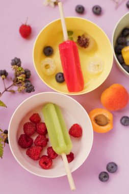 Bright colorful popsicles in colorful bowls with berries and ice cubes on a light violet background. Tasty ice pops close up