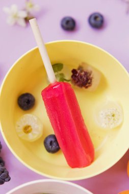 Bright colorful popsicles in colorful bowls with berries and ice cubes on a light violet background. Tasty ice pops close up