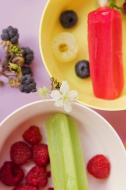Bright colorful popsicles in colorful bowls with berries and ice cubes on a light violet background. Tasty ice pops close up