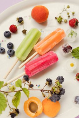 Bright colorful popsicles on a white tray with berries and ice cubes on a light violet background. Tasty ice pops close up