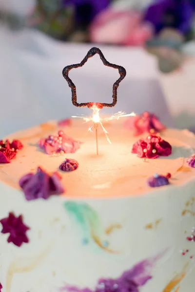 Birthday cake with star sparklers candle on the white cake stand near beautiful bouquet of flowers. Happy birthday gift concept