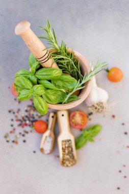 Wooden mortar and pestle with fresh rosemary and basil, spices and tomatoes on a grey concrete background with copy space. Spices and fresh kitchen herbs concept