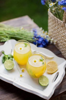 Nane, limon, limon ve portakallı bardaklar saman torbası yanında kır çiçekleriyle birlikte beyaz tahta bir tepside. Yaz içecekleri konsepti
