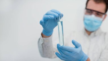 Mavi eldivenli, tıbbi maskeli ve koruyucu gözlüklü beyaz arka planda mavi sıvı maddeli test tüpü tutan bir adam. Tıbbi bayrak konsepti