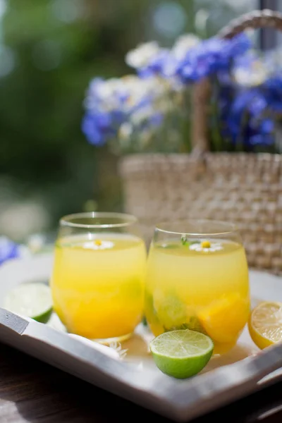 stock image Glasses with lemonade with mint, lime, lemons and orange on a white wooden tray near straw bag with wildflowers. Summer drinks concept