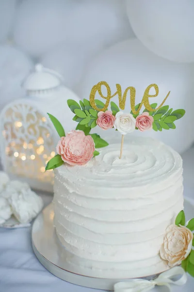 First birthday cake with white cream, golden cake topper with paper flowers. White birthday party for little girl