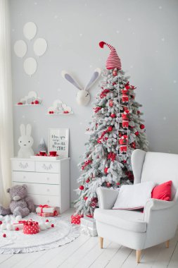 Kırmızı süslemeli Noel ağacı ve gri duvarlı şirin çocuk odasındaki yastıklı beyaz koltuk.
