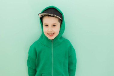 Yeşil kapüşonlu beyaz çocuk açık yeşil arka planda kameraya bakıyor ve gülümsüyor.