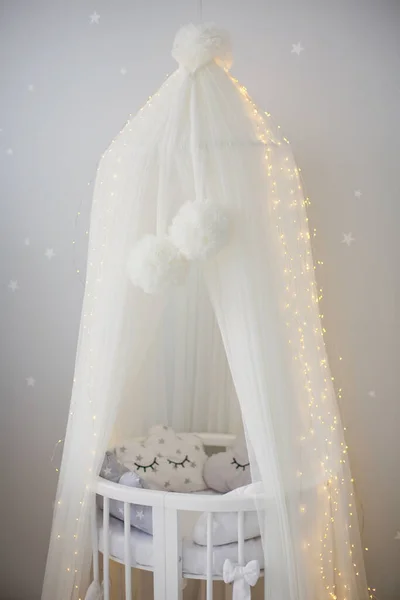 White childs bed crib with canopy and lights near white wall with copy space. Cute infant baby crib with pillows. Light kids interior