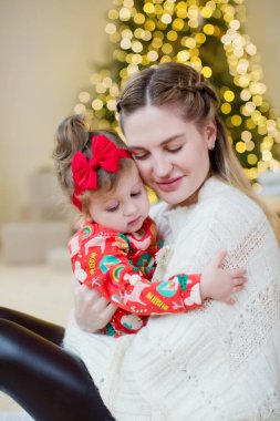 Genç anne ve kızı Noel ağacının yanında. Noel ve yeni yılı bekleyen mutlu bir aile. Noel süslemeleri ve ışıklarla donatılmış rahat bir iç mekan.