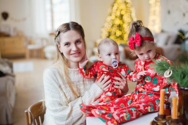 Küçük bir oğlu olan genç bir anne ve kırmızı pijamalı küçük bir kız Noel mutfağında Noel süslemeleri ve ışıklarıyla masada oturuyorlar. Noel arifesinde mutlu bir aile