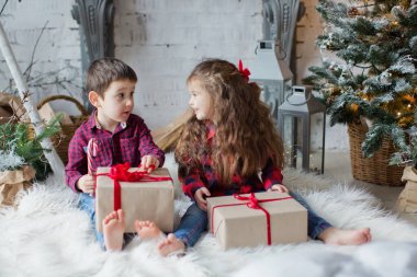 Küçük bir çocuk ve kırmızı gömlekli bir kız Noel ağacının yanında hafif bir Noel arifesinde hediye kutuları tutuyorlar. Tatlı Noel çocukları.
