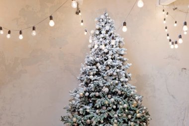 Altın süslemeli ve ışık duvarının yanındaki çelenkli Noel ağacı