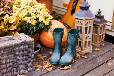 Yeşil çizmeler, ahşap fenerler, hasır sepet, balkabağı ve çiçekler arka bahçede.