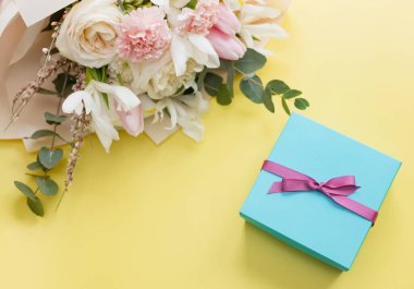 Beautiful bouquet with roses, carnations, iris and eucalyptus and blue gift box with pink ribbon on a yellow background, copy space. Mothers day, 8 march, holiday gift concept