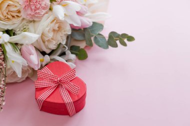 Beautiful bouquet with roses, carnations, iris and eucalyptus and red heart gift box on a pink background, copy space. Mothers day, 8 march, holiday gift concept