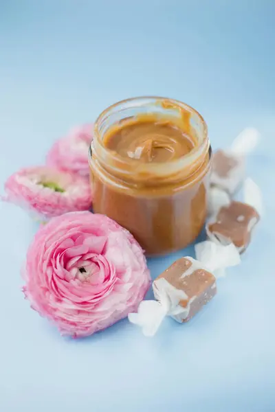 Jar with caramel sauce, caramel candies in paper wrapping, pink ranunculus flowers on a blue background with copy space