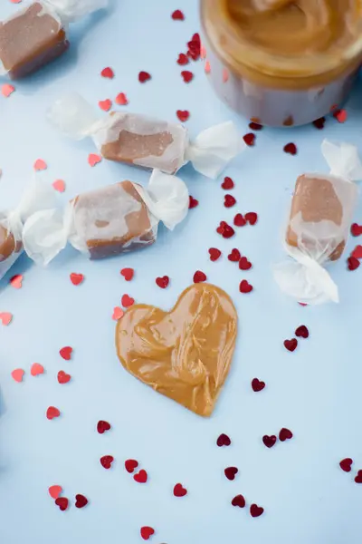Jar with caramel sauce, caramel candies in paper wrapping, heart shaped caramel decoration on a blue background with red hearts confetti