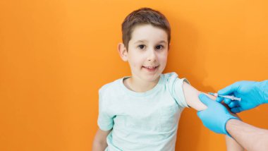 Aşı konsepti. Mavi eldivenli doktor, turuncu arka planda mavi tişörtlü ve fotokopi alanı olan beyaz çocuğu aşılıyor.
