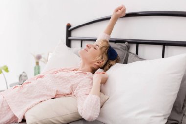 Pembe pijamalı ve uyku maskeli bir kadın parlak bir yatak odasında uyanır.