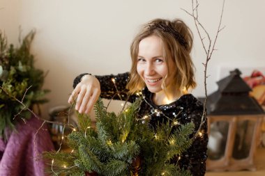 Siyah elbiseli ve açık renk çelenkli bir kadın Noel süslerinin ve mumlu fenerin yanında oturuyor.