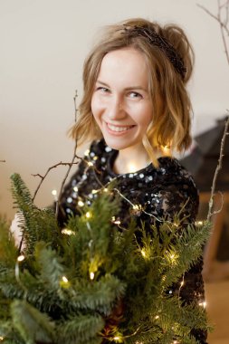 Siyah elbiseli ve açık renk çelenkli bir kadın Noel süslerinin ve mumlu fenerin yanında oturuyor.