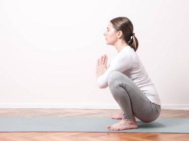 Kahverengi saçlı beyaz kadın kurbağa duruşunda yoga yapıyor ya da Malasana soldan kameraya mavi gri hasırda poz veriyor ve ahşap zemin iç tarafta beyaz duvar arka planı olan bir iç oda.. 