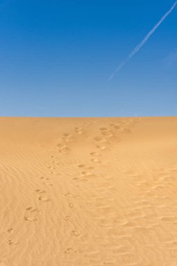 Arka planda mavi gökyüzü olan kum tepelerinde ayak izleri var.