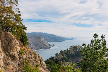 Bulutlu bir gökyüzünün altında, kayalık dağlardan görünen mavi okyanus.