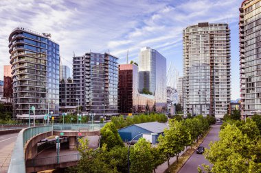 Vancouver, Kanada - 4 Haziran 2023 - Çağdaş yüksek binalar, yemyeşil ağaçlar ve mavi gökyüzünün altında kıvrımlı bir üst geçidin yer aldığı canlı bir şehir manzarası. Sahne, şehir mimarisi ve doğal elementlerin bu 'daki karışımını yakalıyor.