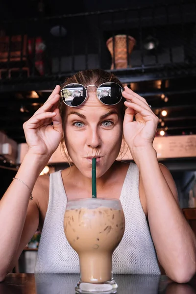 ストローでアイスコーヒーを飲みながらサングラスを保持している面白い女性の垂直写真 — ストック写真