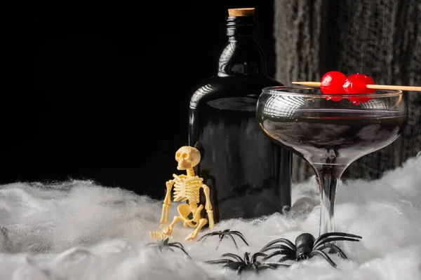 stock image close-up of black Halloween cocktail glass with red cherries, skeletons, spiders and dark bottle on spider web, black background, horizontal, with copy space