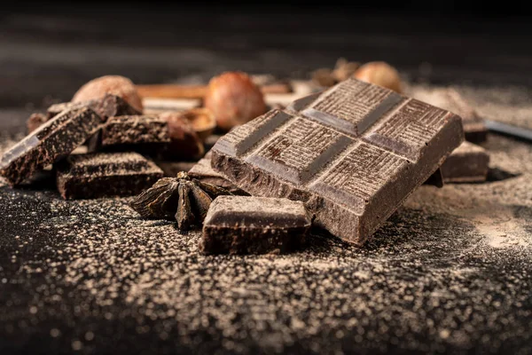 Close-up of pieces of chocolate with cocoa powder and hazelnuts on dark table, black background, horizontal, with copy space