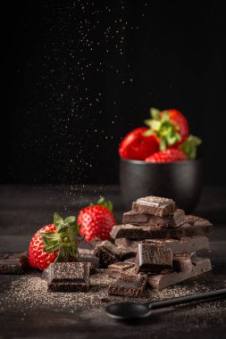 Çikolata parçalarının üzerine dökülen kakaonun görüntüsü kasede çilek ve koyu ahşap masa üzerinde kaşık, siyah arka plan, dikey, kopyalama alanı olan