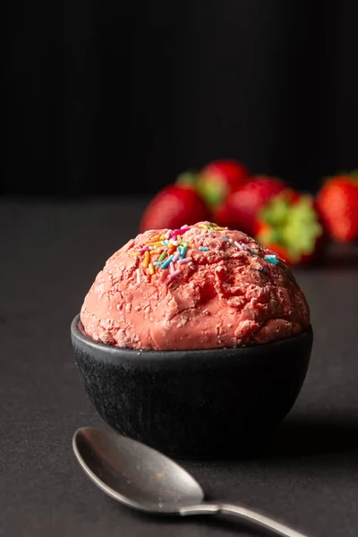 Close up of strawberry ice cream in bowl and black table with spoon and strawberries, black background, vertical, with copy space