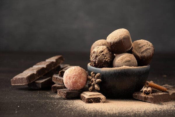 View of chocolates with cocoa powder in bowl on wooden table with pieces of chocolate, cinnamon and anise, selective focus, gray background, horizontal, with copy space