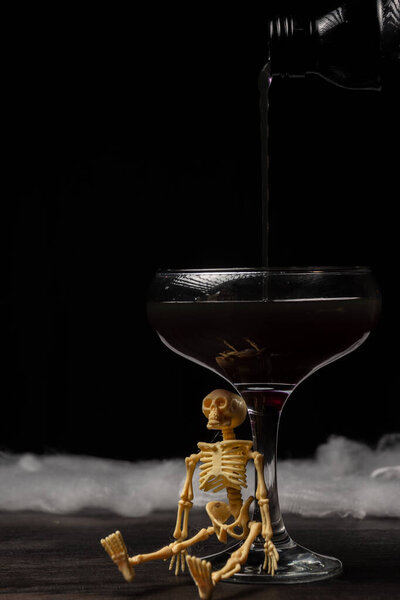 View of bottle pouring black Halloween cocktail into glass with skeleton on table and black background, vertical, with copy sapce