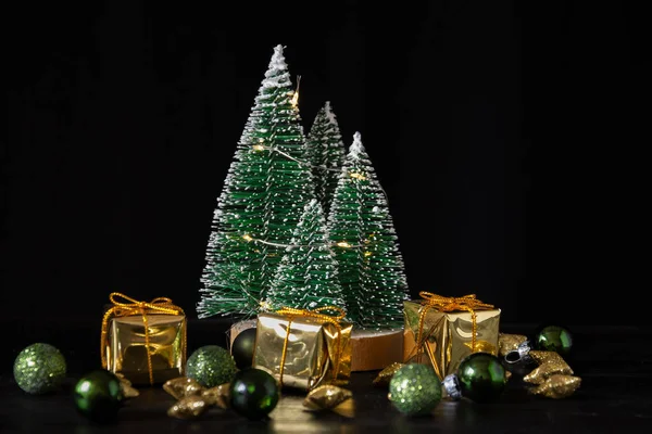 close-up of little trees with lights on dark table with Christmas decorations in green and gold, black background, horizontal, with copy space