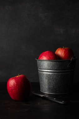 Koyu ahşap masa üzerinde kırmızı elmalı metal kovanın görüntüsü. Bıçak ve elmalı, siyah arka plan, dikey, kopyalama alanı olan.