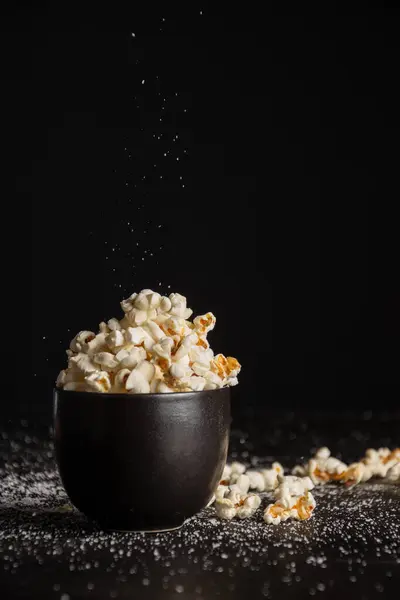 View of black bowl with popcorn on dark table with salt , selective focus, black background, vertical, with copy space