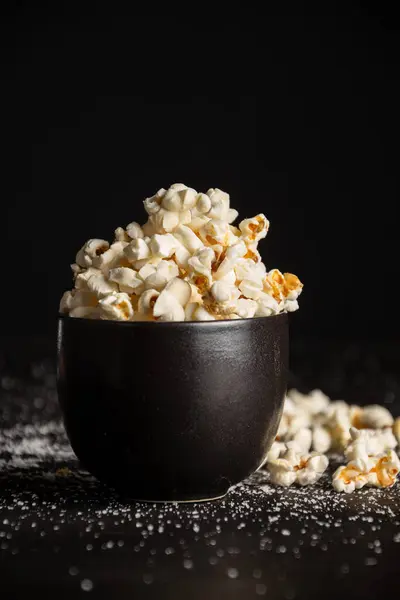 Closeup of black bowl with popcorn on dark table with salt , selective focus, black background, vertical, with copy space