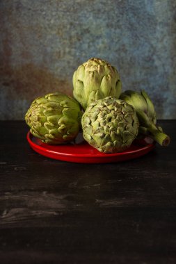 Koyu ahşap masa üzerinde dört enginar bulunan kırmızı plaka görüntüsü, gri arkaplan, dikey, kopyalama alanı ile