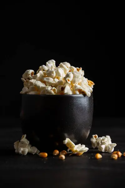 Close-up of black bowl with popcorn on dark table with popcorn, grains and salt, black background, vertical, with copy space